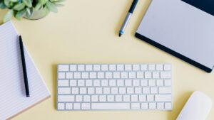 White keyboard, notebooks and pens arranged on a yellow desk.
